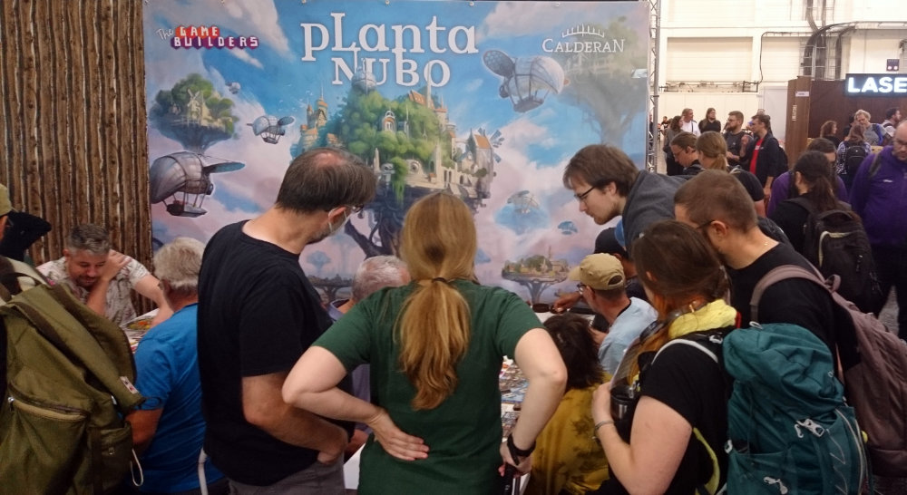 Menschen stehen und sitzen um einen Planta-Nubo-Spieltisch auf der Spiel ’23 in Essen. Im Hintergrund ein großes Banner, welches den Planta-Nubo-Titel zeigt. Eine Illustration im Studio-Ghibli-Style, welches einen riesigen Baum zeigt, auf dem eine grüne Stadt gebaut ist.